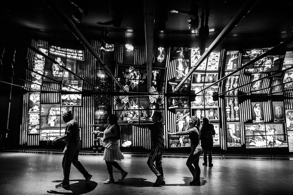 Des silhouettes en mouvement devant la mosaïque lumineuse de la Place des Arts, en contre-jour et en noir et blanc.