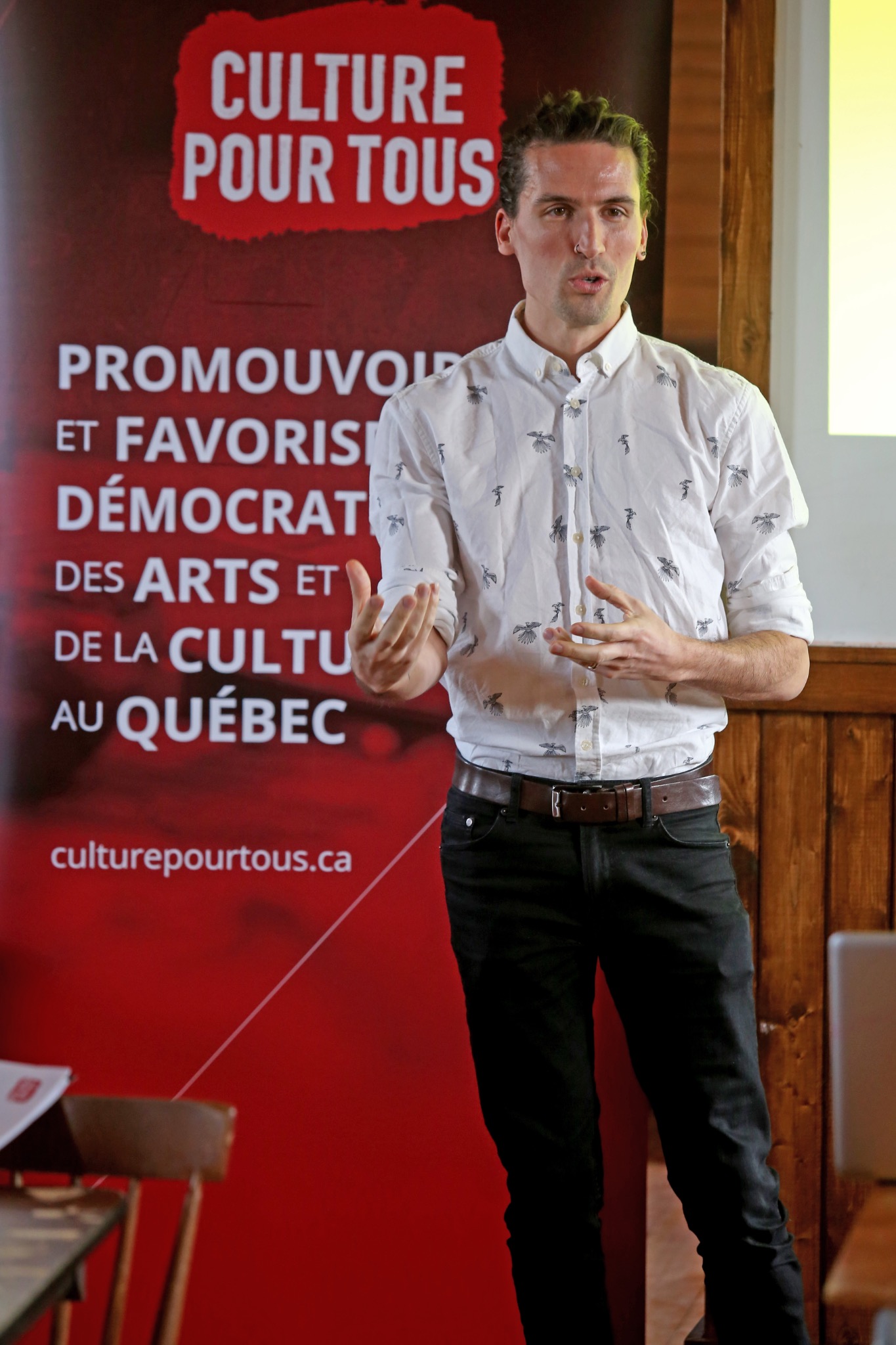 Marco Pronovost donne une conférence devant une bannière Culture pour tous, en chemise blanche à petits motifs noirs.